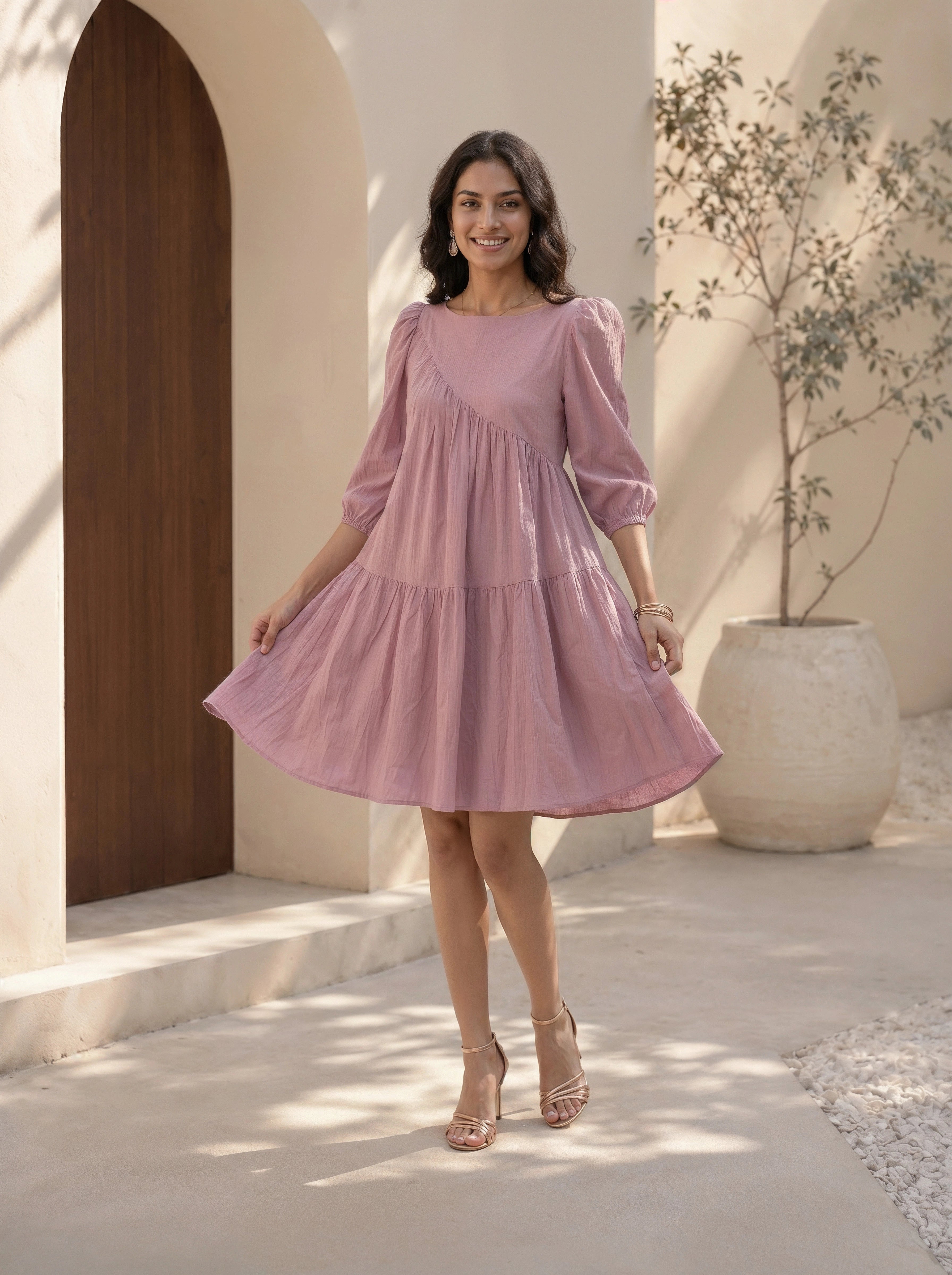 Woman in a pink dress standing in a sunlit outdoor setting with a wooden door and potted plant in the background.
