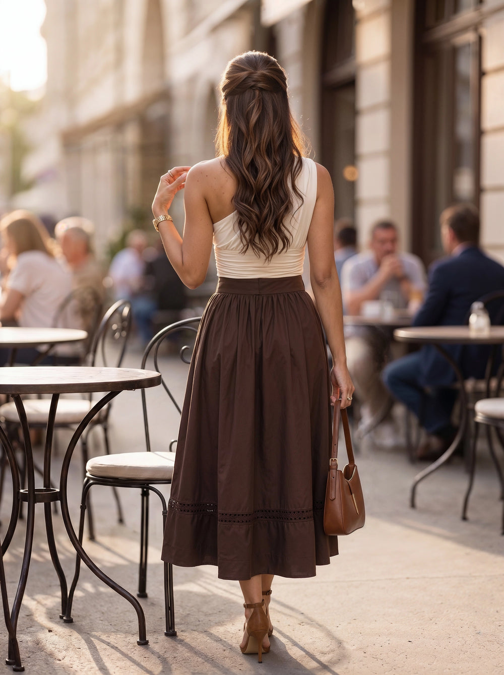 A woman viewed from behind walking on a city sidewalk past outdoor cafe tables, wearing a sleeveless cream top and a long brown midi skirt, with a brown handbag and tan heeled sandals.