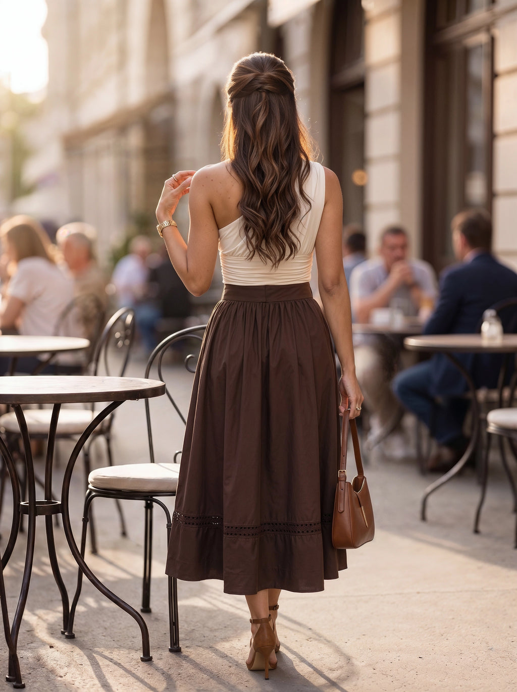 A woman viewed from behind walking on a city sidewalk past outdoor cafe tables, wearing a sleeveless cream top and a long brown midi skirt, with a brown handbag and tan heeled sandals.