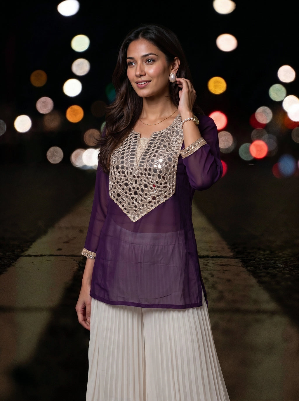 A woman wearing a purple sheer kurta with a beige crochet yoke and white pleated pants, standing on a city street at night with blurred lights in the background.