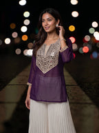 A woman wearing a purple sheer kurta with a beige crochet yoke and white pleated pants, standing on a city street at night with blurred lights in the background.