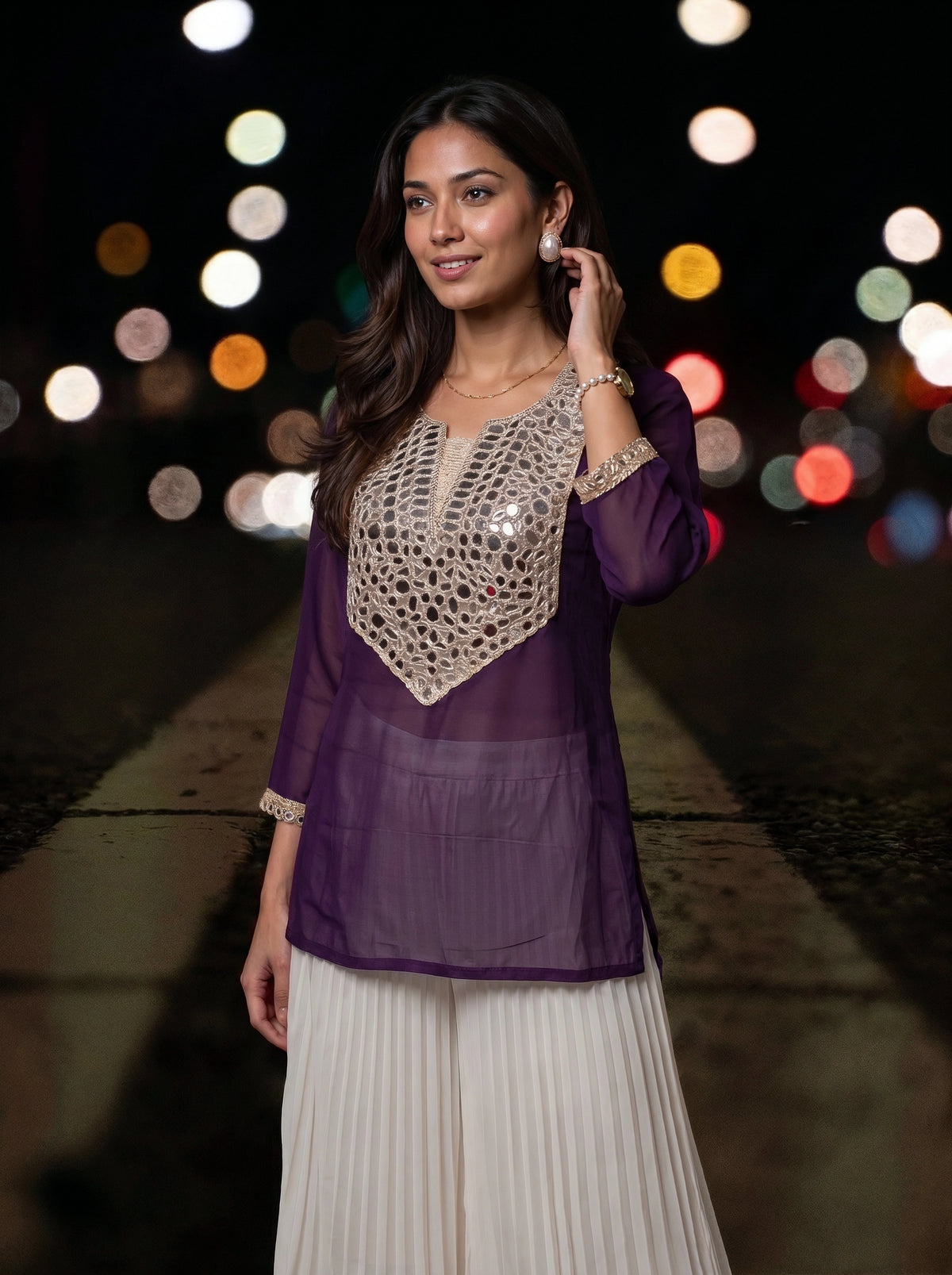 A woman wearing a purple sheer kurta with a beige crochet yoke and white pleated pants, standing on a city street at night with blurred lights in the background.