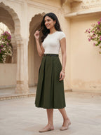 Woman wearing a white top and green skirt standing in front of an architectural background with flowers.
