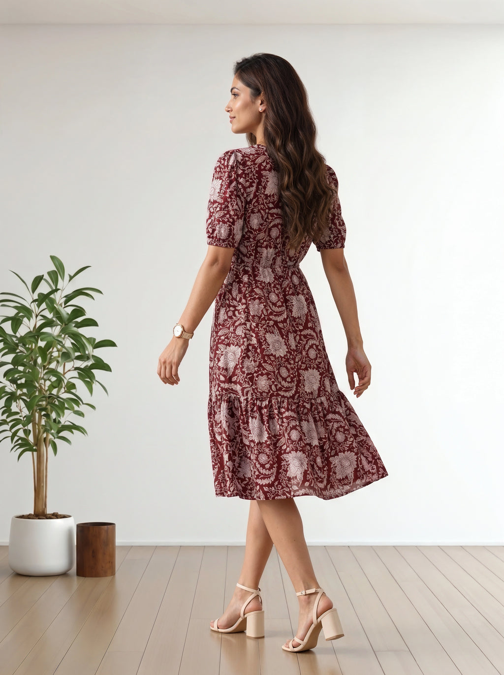 A woman wearing a short-sleeved midi dress with a red and beige floral paisley pattern, shown from the back in a bright room with a wooden floor and a potted plant.
