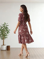A woman wearing a short-sleeved midi dress with a red and beige floral paisley pattern, shown from the back in a bright room with a wooden floor and a potted plant.