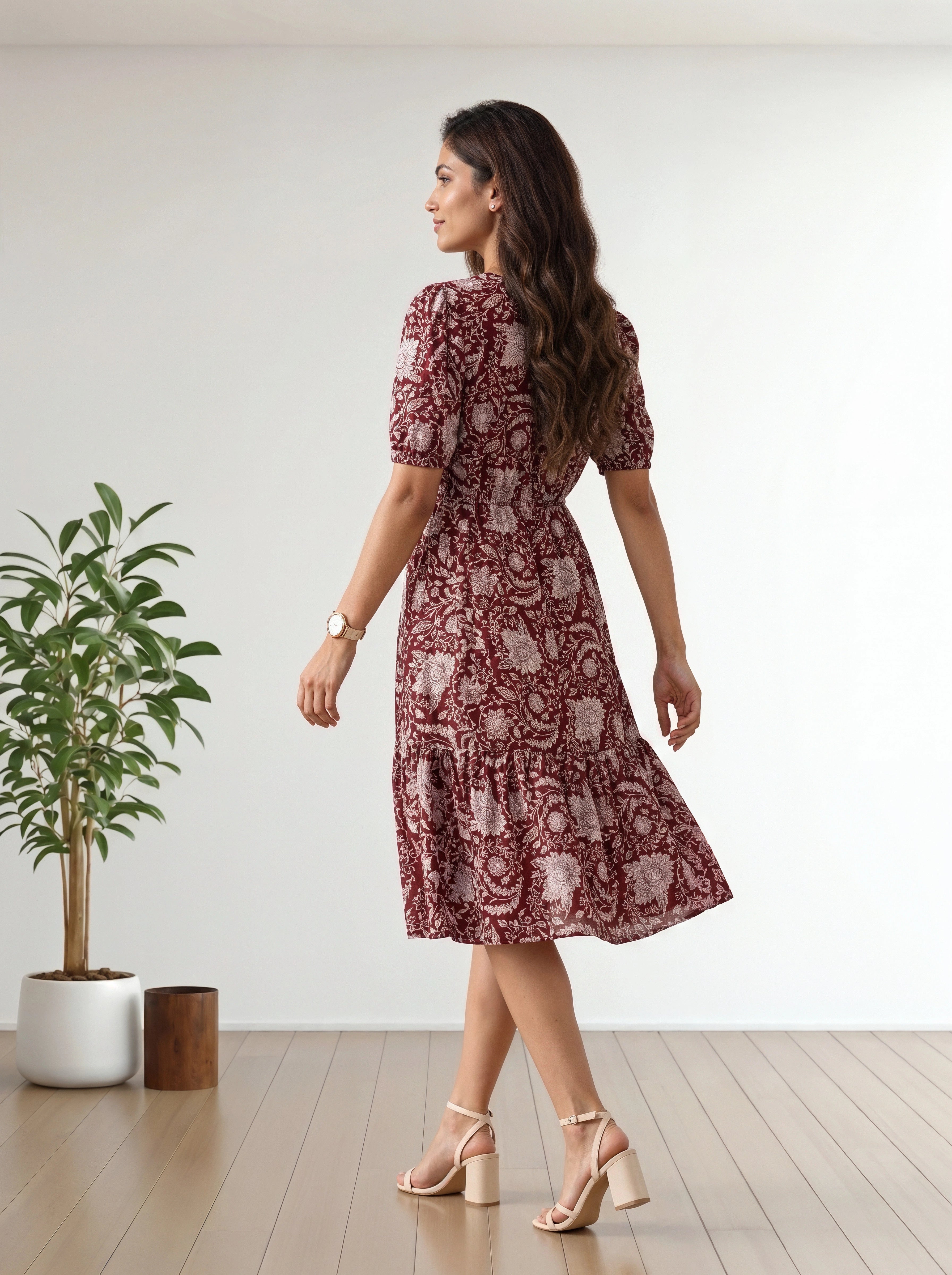 A woman wearing a short-sleeved midi dress with a red and beige floral paisley pattern, shown from the back in a bright room with a wooden floor and a potted plant.
