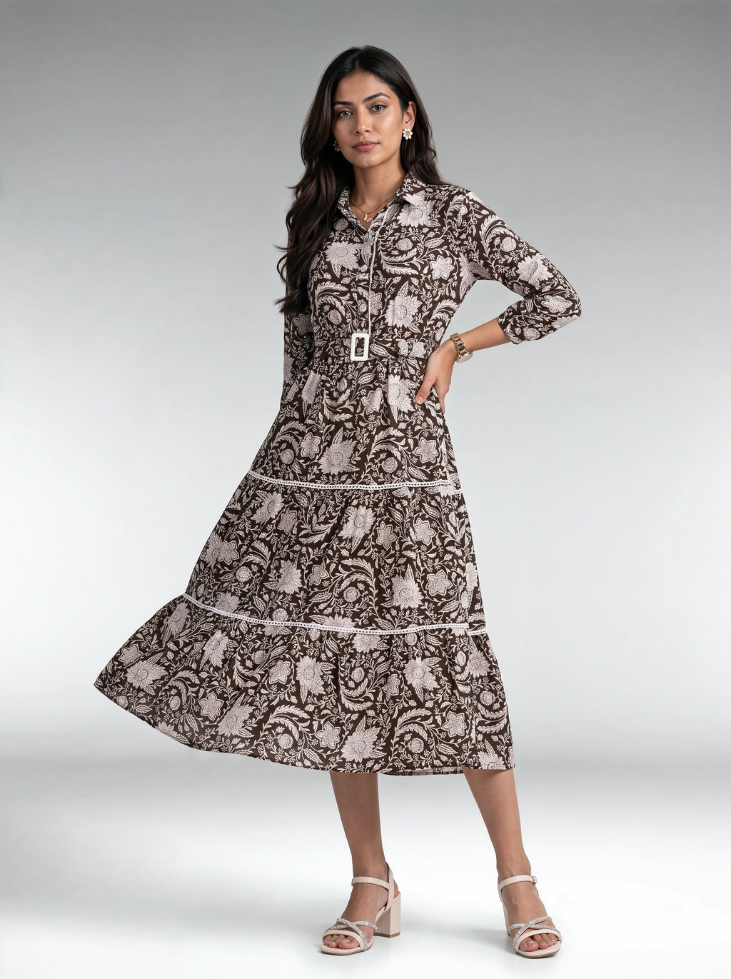 A woman modeling a long-sleeve midi dress with a brown and beige paisley pattern, featuring a matching belt with a silver buckle, set against a neutral gray studio background.