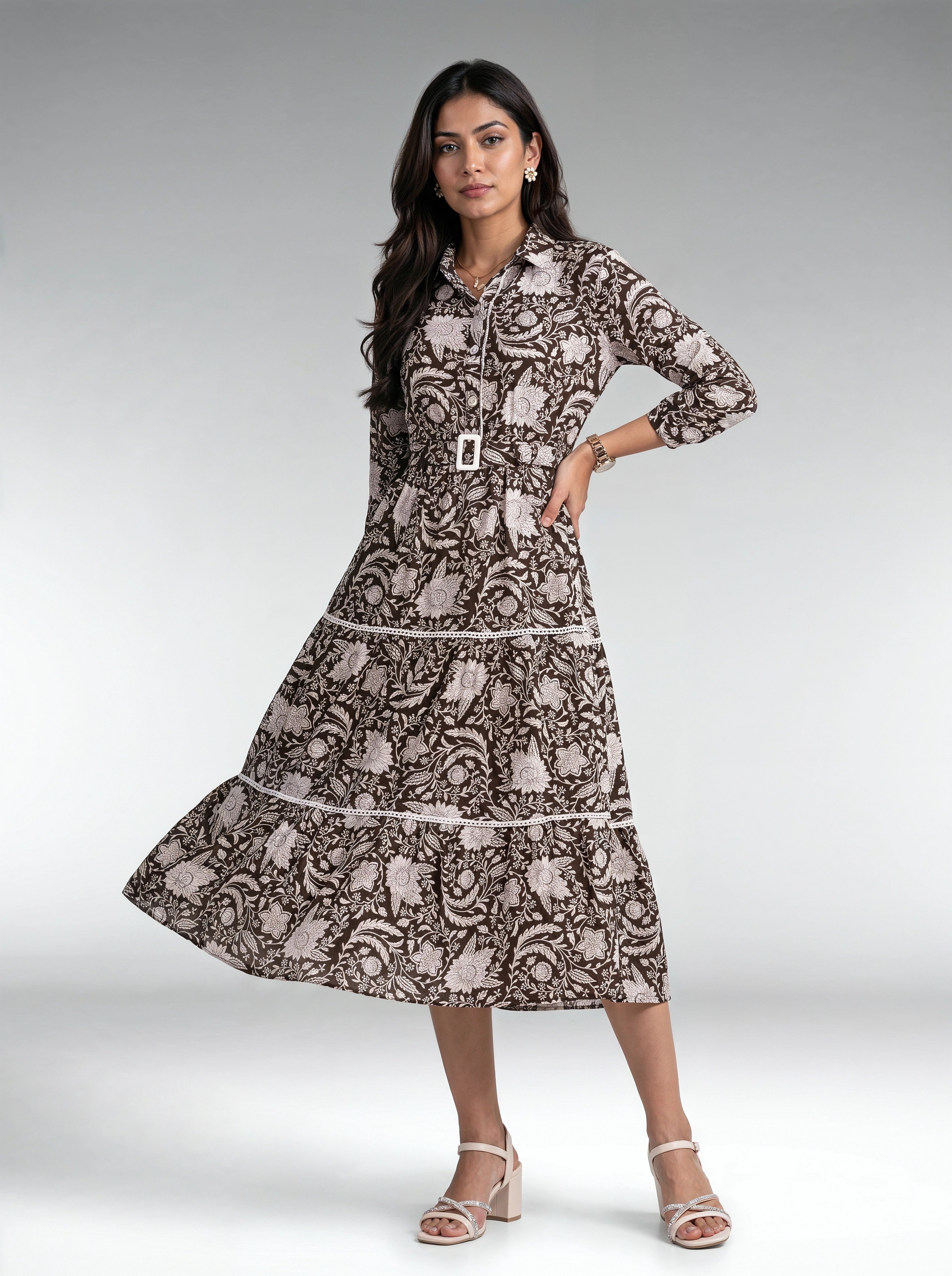 A woman modeling a long-sleeve midi dress with a brown and beige paisley pattern, featuring a matching belt with a silver buckle, set against a neutral gray studio background.