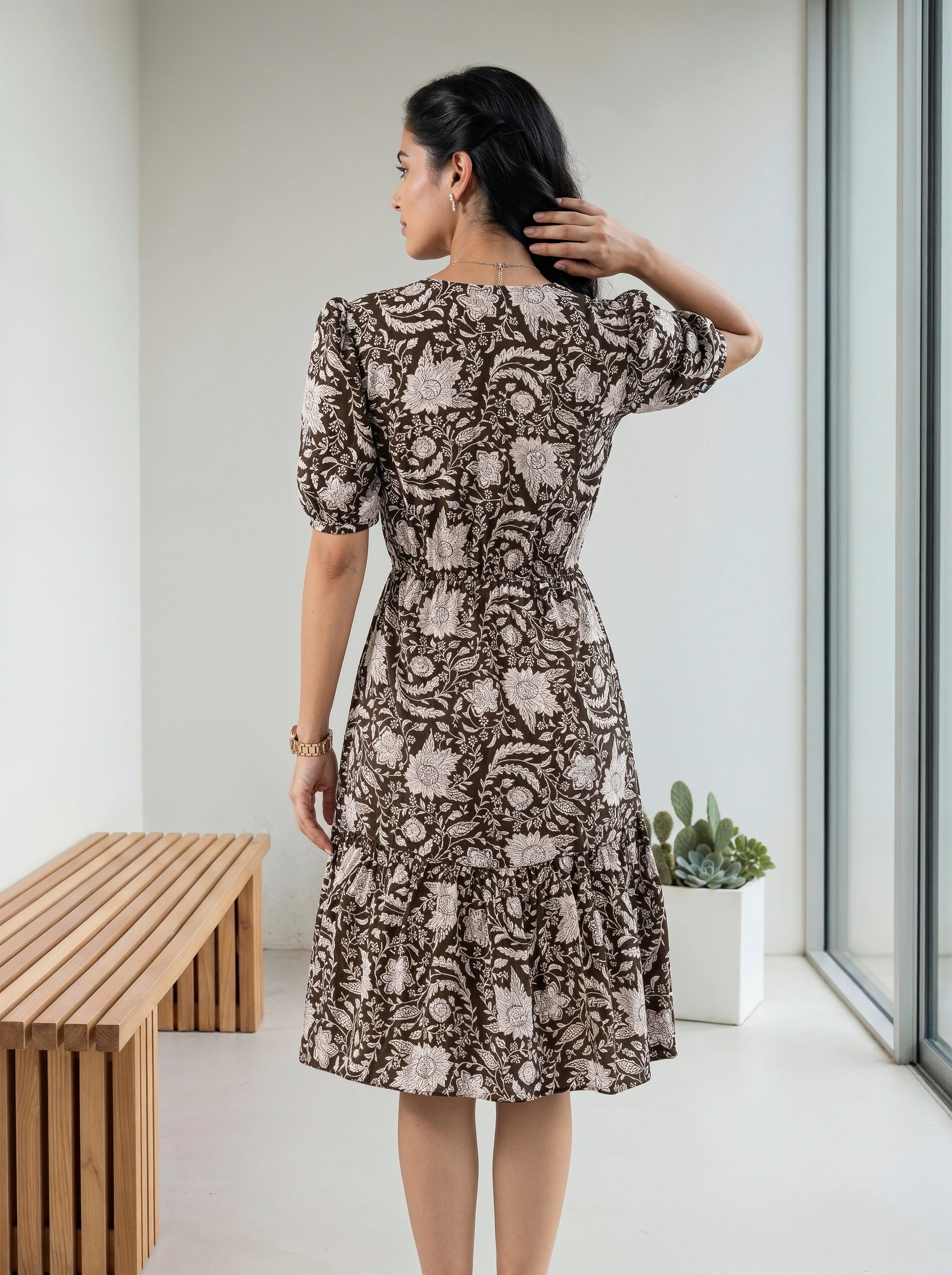 A woman wearing a brown and beige floral print dress, shown from the back in a bright indoor setting with a wooden bench and window.