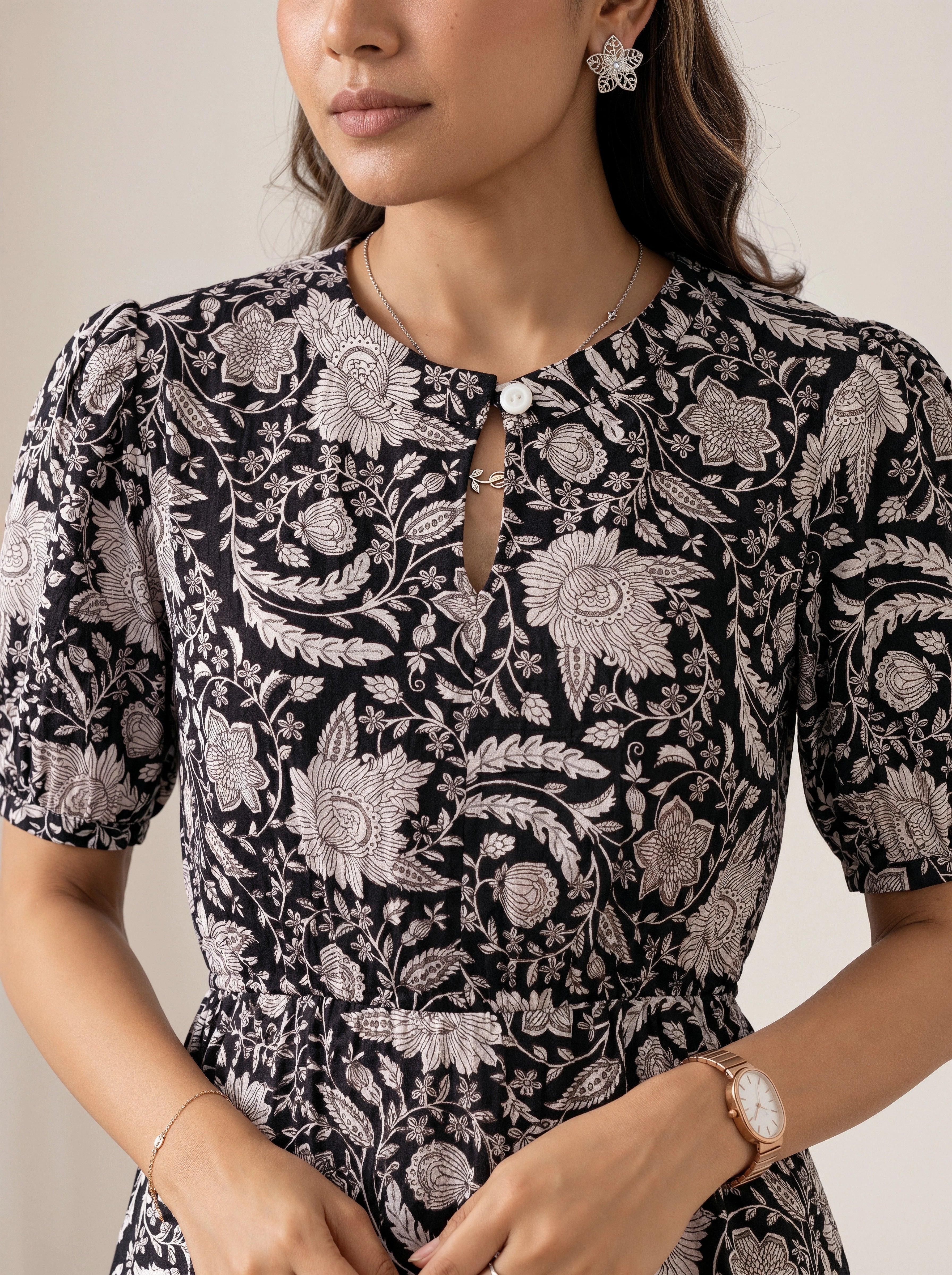 A close-up of a woman wearing a short-sleeve black dress with a detailed white floral and botanical pattern, featuring a keyhole neckline with a decorative white bead and keyhole closure.