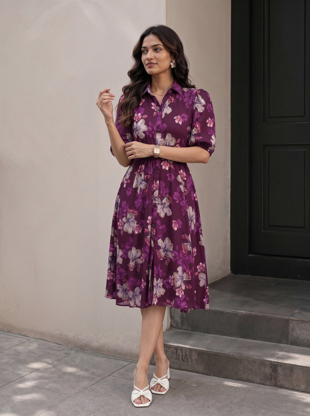 A woman wearing a black midi dress with three-quarter sleeves, a tie-neck detail, and a gathered waist with a matching fabric tie, shown in a warm-toned indoor setting.