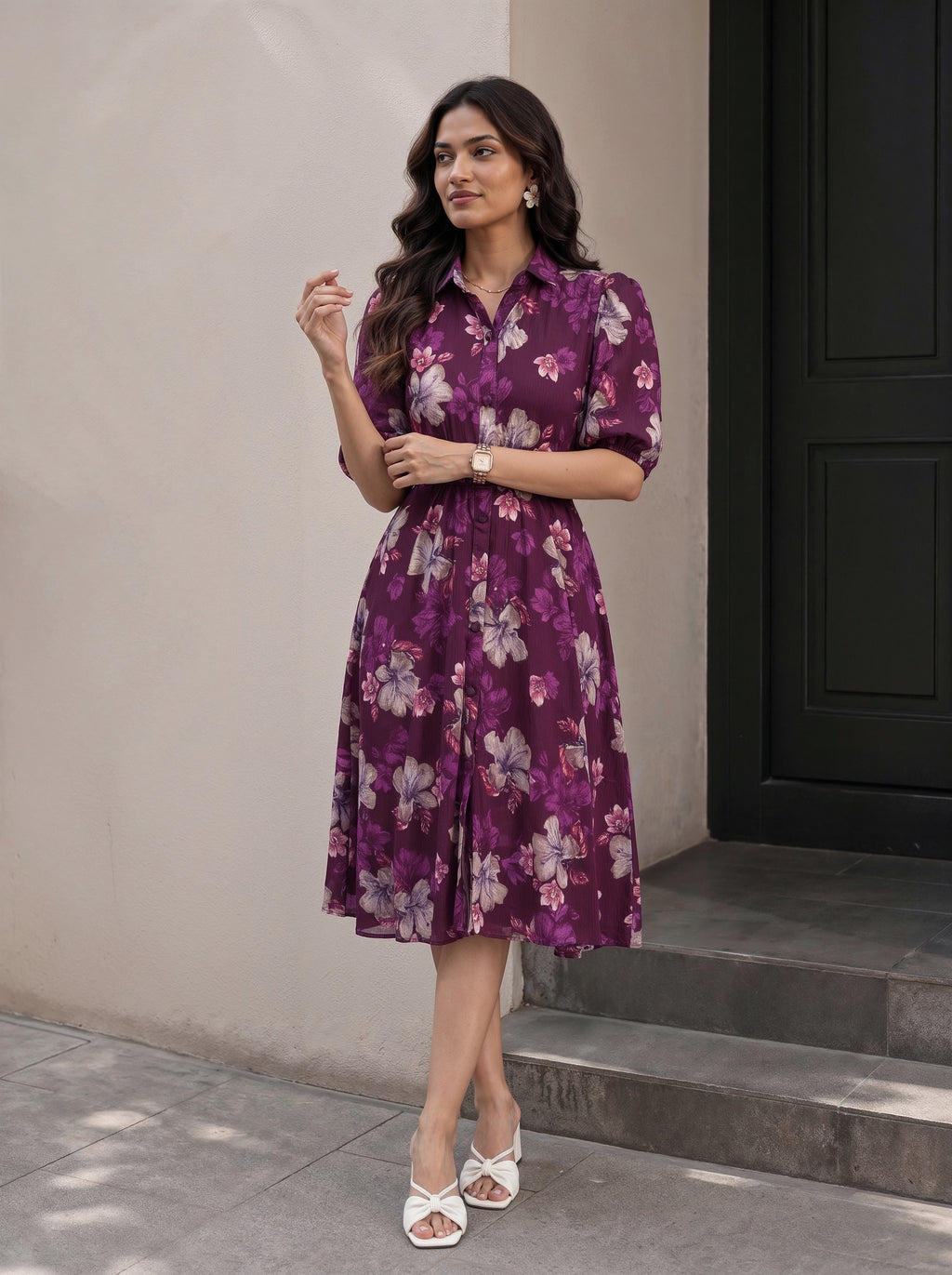 A woman wearing a black midi dress with three-quarter sleeves, a tie-neck detail, and a gathered waist with a matching fabric tie, shown in a warm-toned indoor setting.