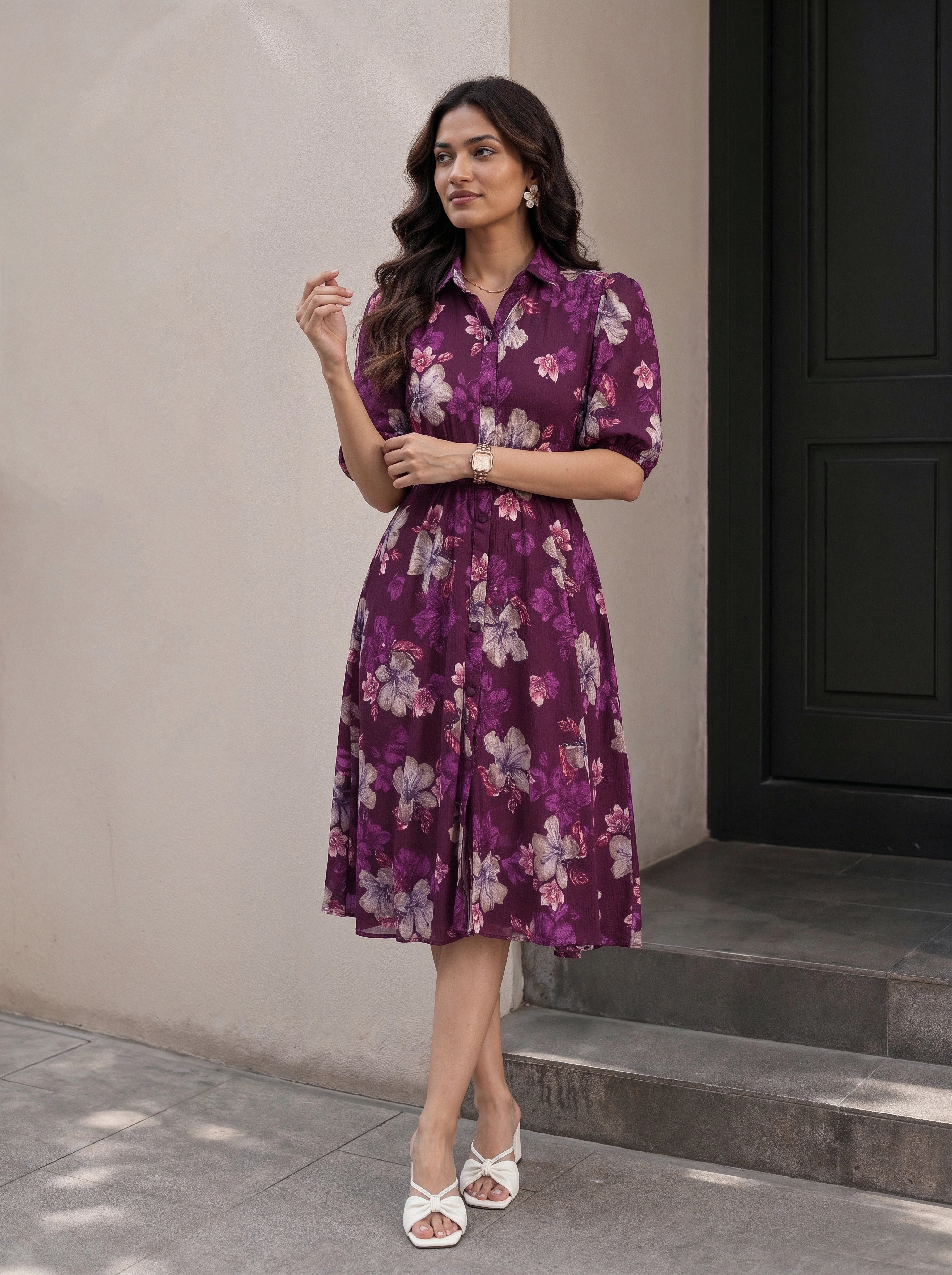 A woman wearing a black midi dress with three-quarter sleeves, a tie-neck detail, and a gathered waist with a matching fabric tie, shown in a warm-toned indoor setting.