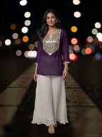 A woman modeling a traditional Indian outfit consisting of a purple kurta with a heavily embellished silver sequin V-neck bodice and white pleated palazzo pants, set against a dark background with colorful bokeh lights.