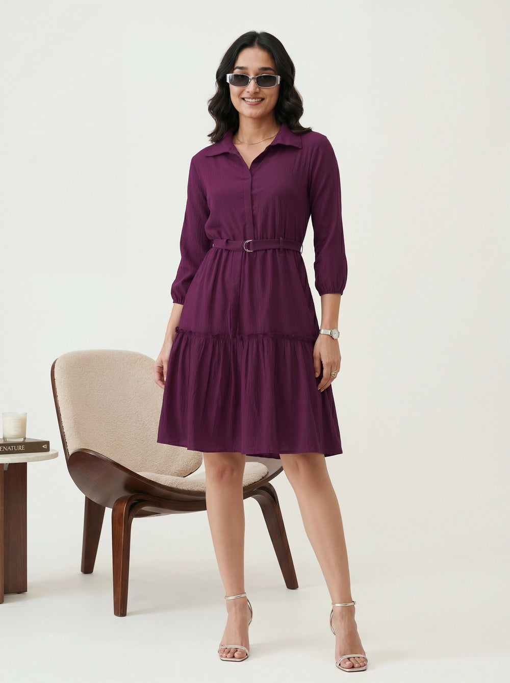 Woman wearing a purple dress standing in a minimalistic room with a chair and table.