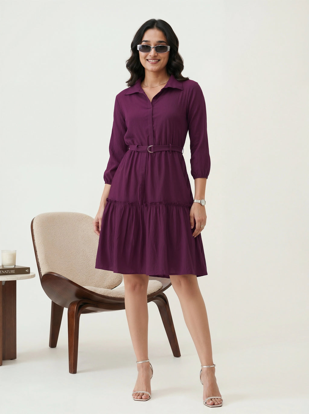 Woman wearing a purple dress standing in a minimalistic room with a chair and table.