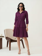 Woman wearing a purple dress standing in a minimalistic room with a chair and table.