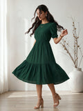 Woman wearing a green dress standing in a bright room with a vase in the background