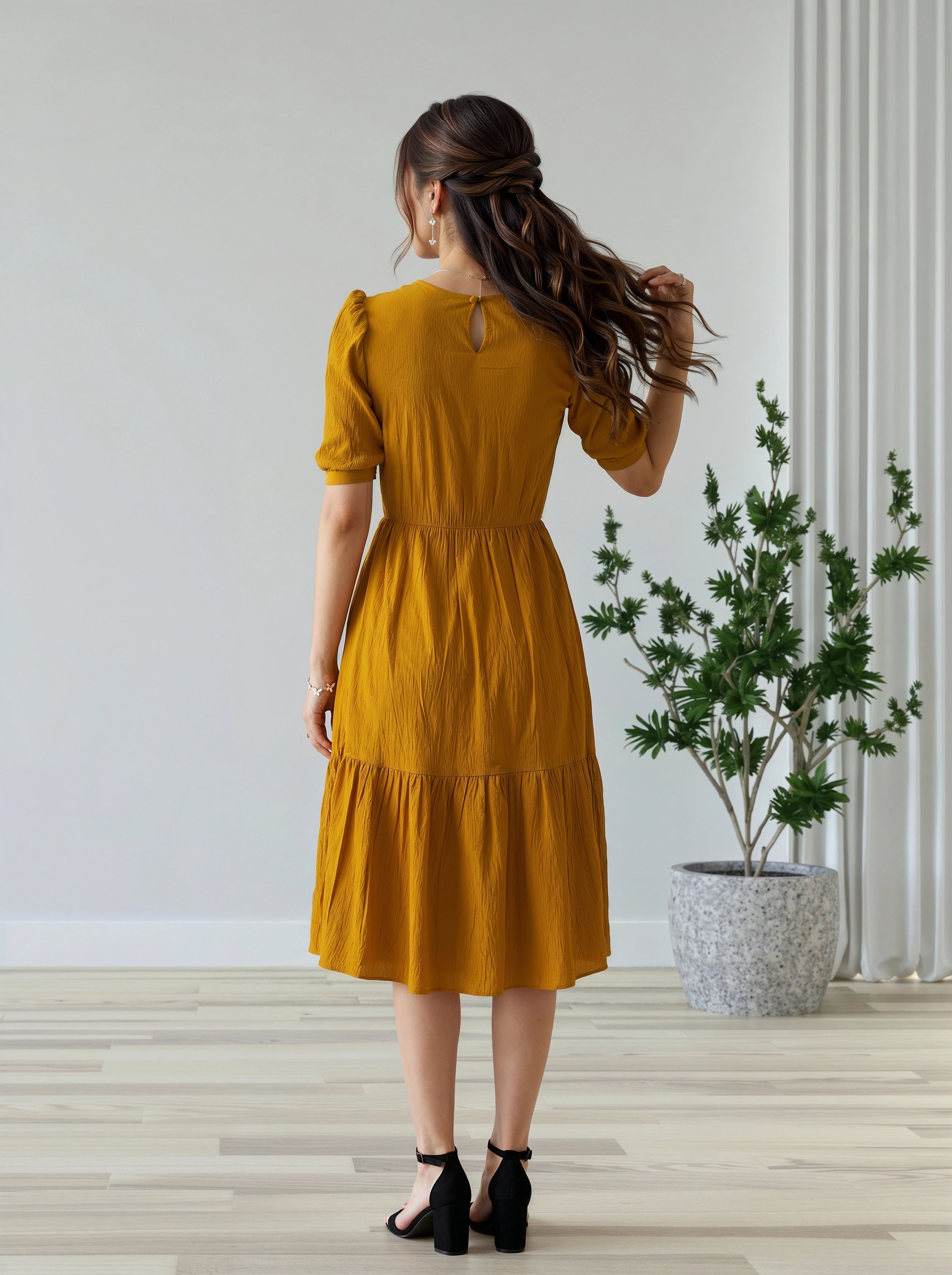 Woman in a mustard yellow dress standing in a room with a plant and white wall.