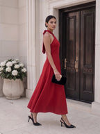 A woman models a sleeveless red midi dress with a tie-neck detail, paired with black heels and a black clutch, standing in front of a grand dark wooden door.