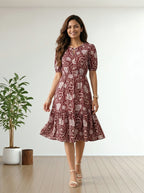 A woman modeling a short-sleeve midi dress with a red and beige paisley pattern, standing in a bright room with a wooden floor and a potted plant.