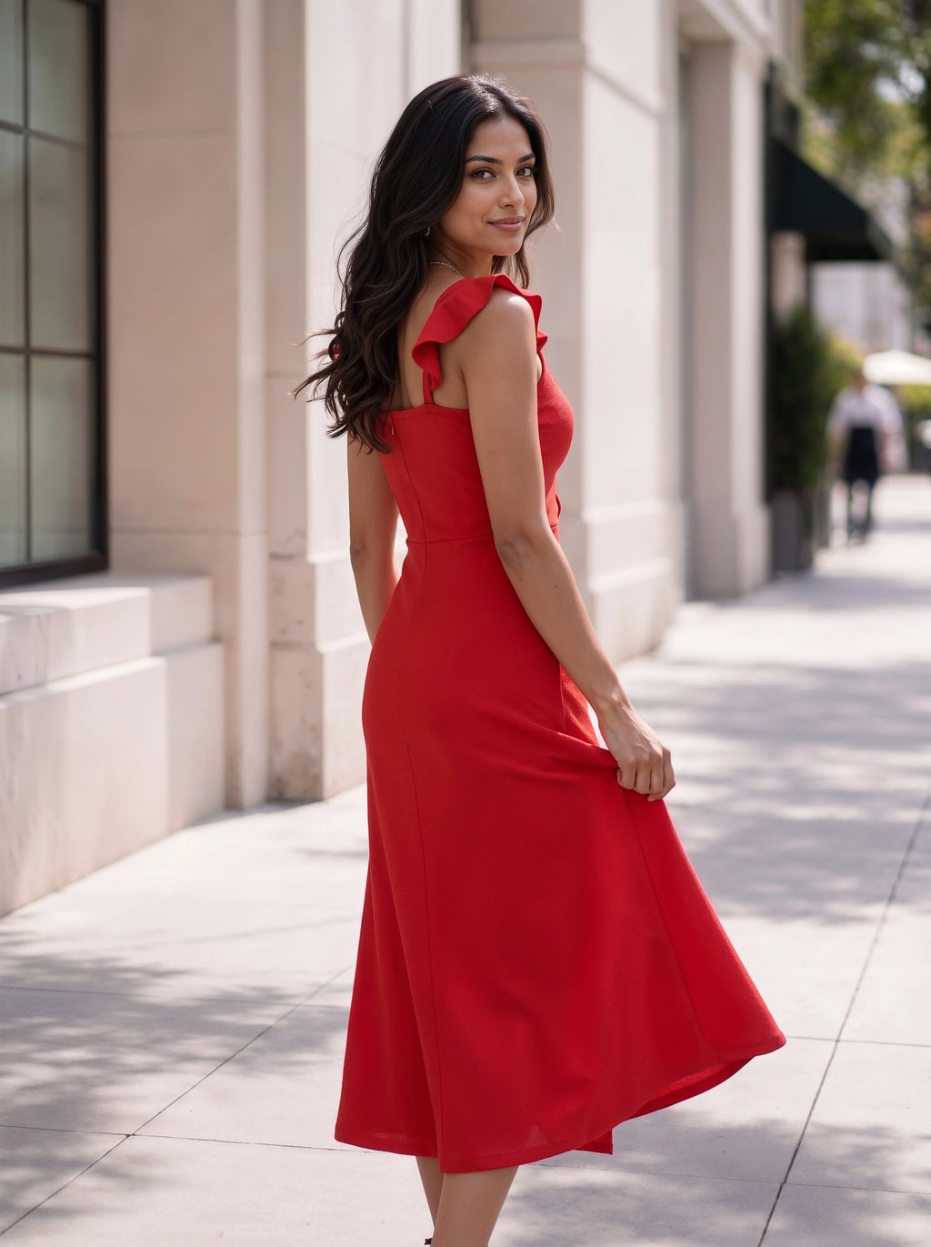 A woman wearing a bright red sleeveless midi dress with ruffled shoulder straps stands on a sunlit city sidewalk, holding out one side of the skirt to show its flow.