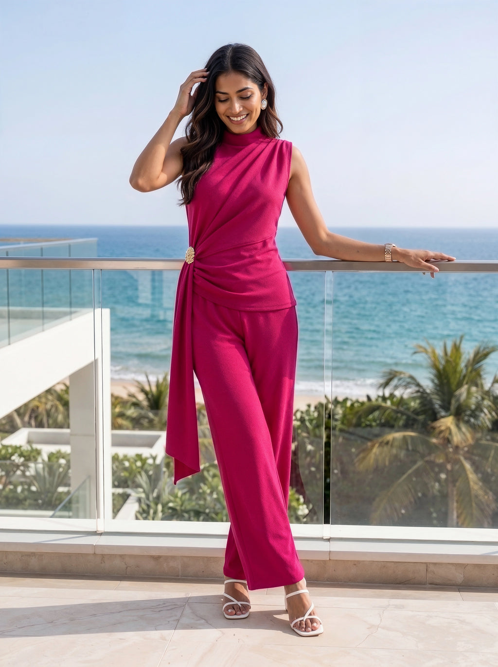 A woman modeling a bright magenta pink sleeveless wrap-style jumpsuit with a gold button accent at the waist, standing on a modern balcony with a tropical ocean and palm tree view in the background.