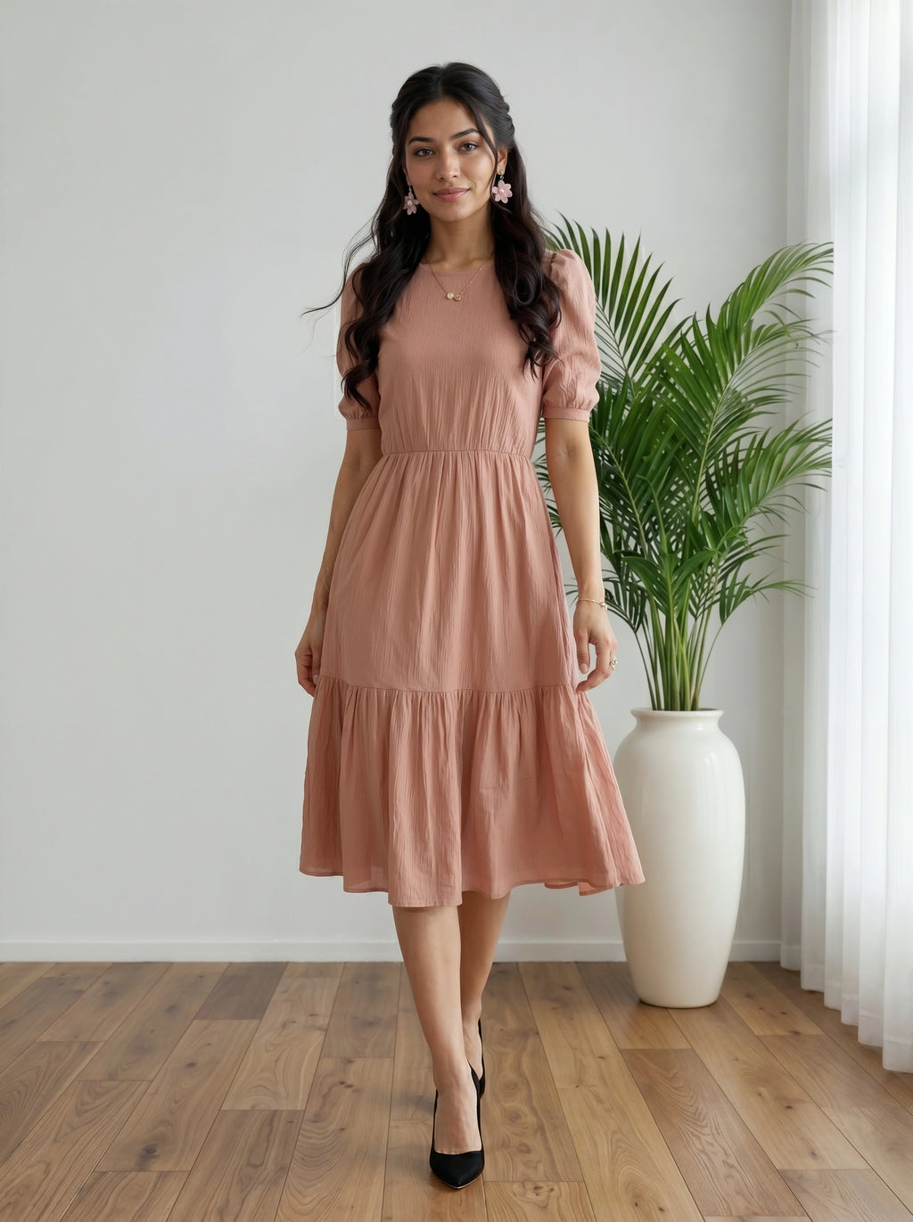 Woman in a pink dress standing in a room with a plant and white curtain.