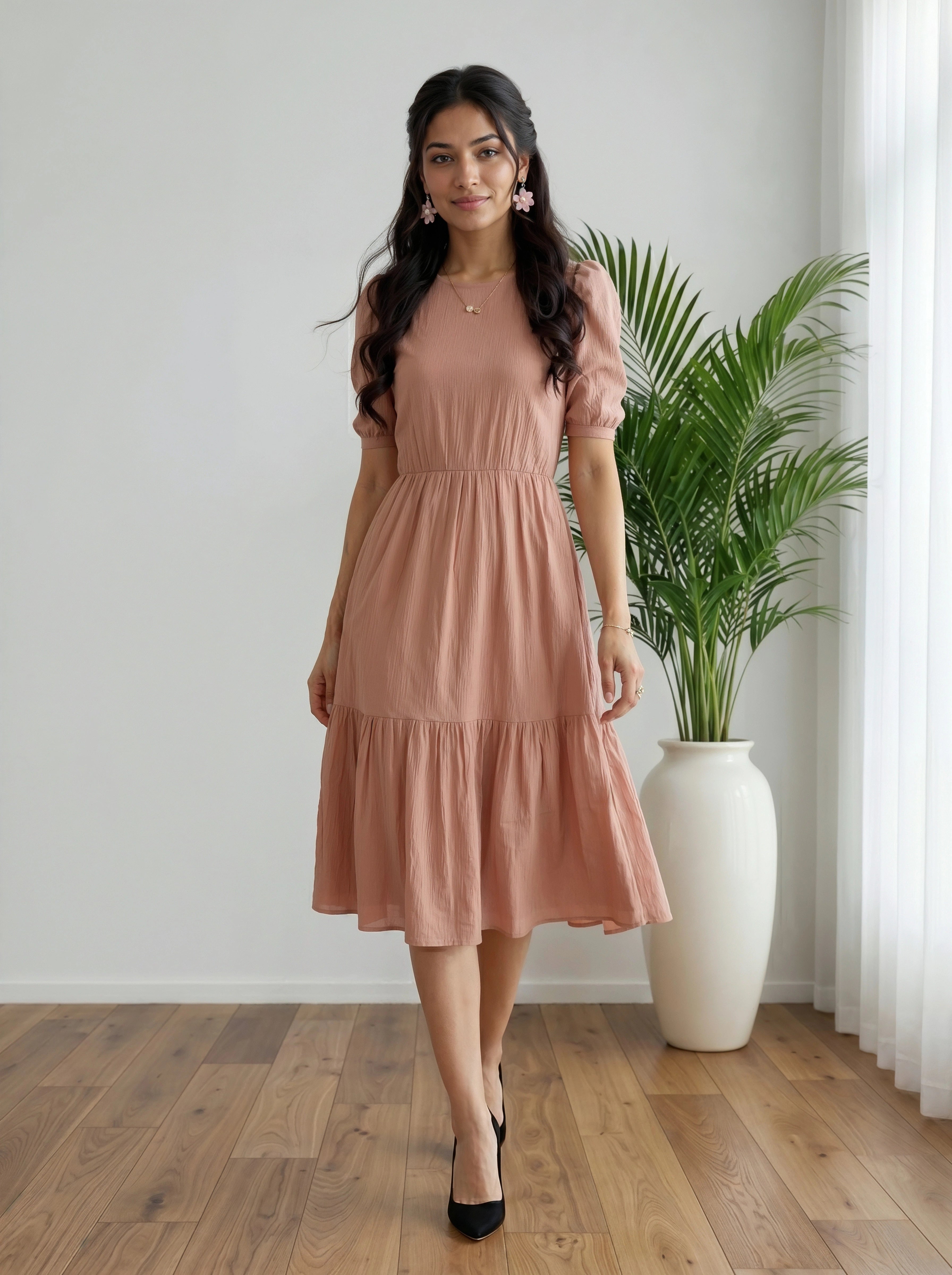 Woman in a pink dress standing in a room with a plant and white curtain.