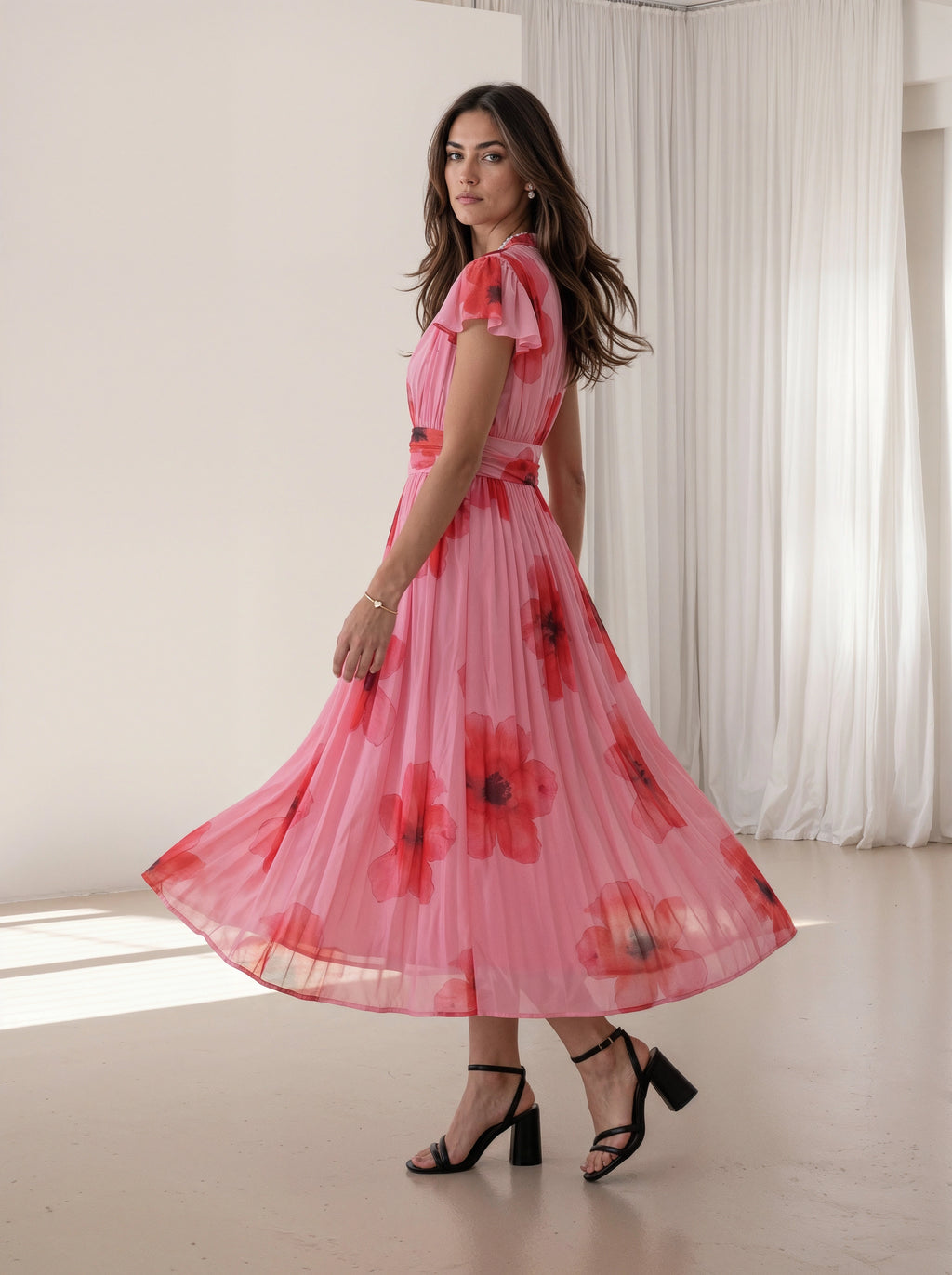 A woman wearing a pink pleated midi dress with a red and black floral print, ruffled cap sleeves, and a matching waist tie, posing in a bright indoor studio with white curtains.