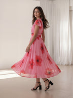 A woman wearing a pink pleated midi dress with a red and black floral print, ruffled cap sleeves, and a matching waist tie, posing in a bright indoor studio with white curtains.