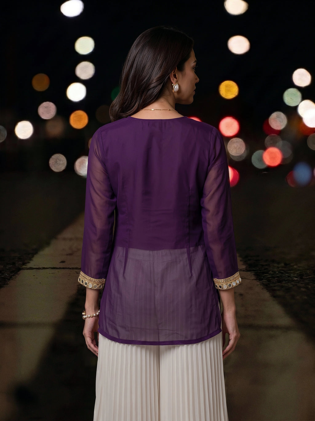 A woman viewed from behind wearing a purple long-sleeve kurta with sheer mesh panels and gold-trimmed cuffs, paired with white pleated pants, set against a blurred night street background with colorful bokeh lights.