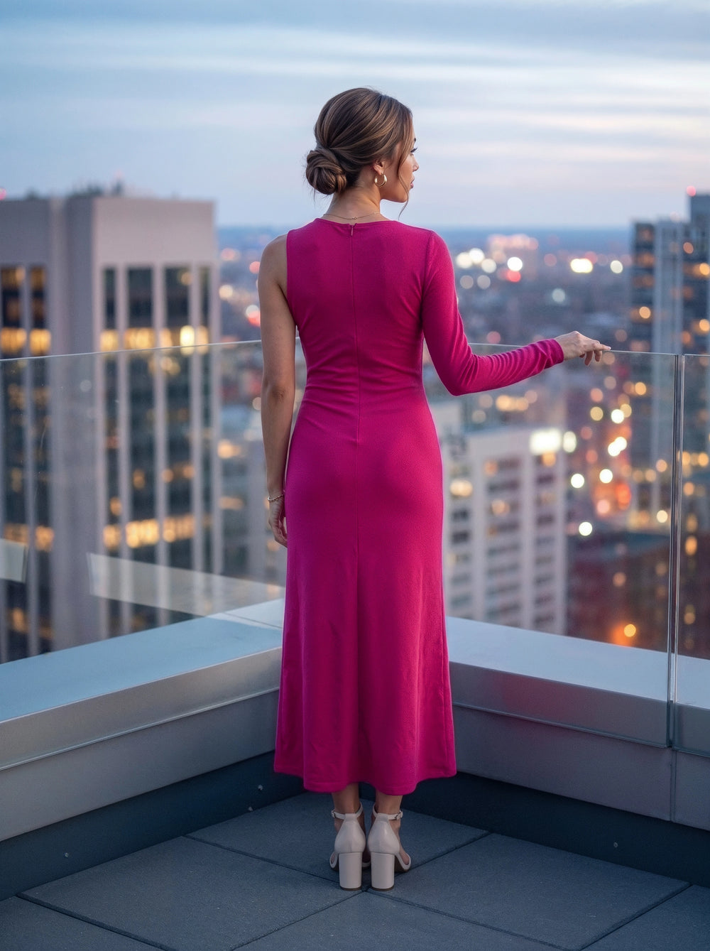 A woman stands on a rooftop terrace at dusk, viewed from behind, wearing a sleeveless magenta-pink long-sleeve maxi dress and white block-heel sandals, with a blurred city skyline in the background.