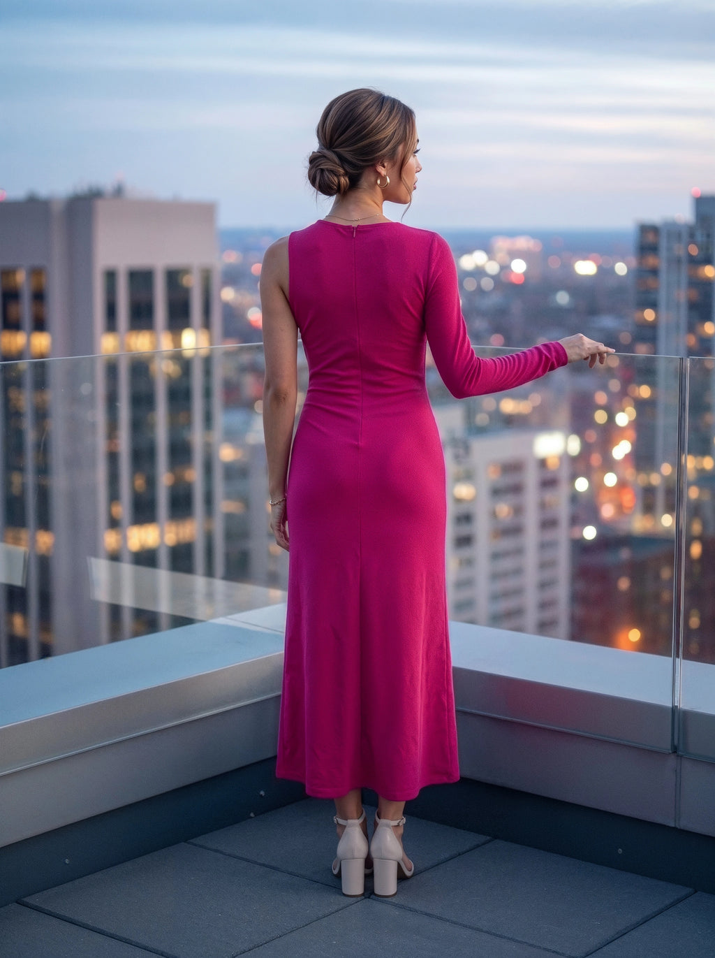 A woman stands on a rooftop terrace at dusk, viewed from behind, wearing a sleeveless magenta-pink long-sleeve maxi dress and white block-heel sandals, with a blurred city skyline in the background.