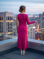 A woman stands on a rooftop terrace at dusk, viewed from behind, wearing a sleeveless magenta-pink long-sleeve maxi dress and white block-heel sandals, with a blurred city skyline in the background.