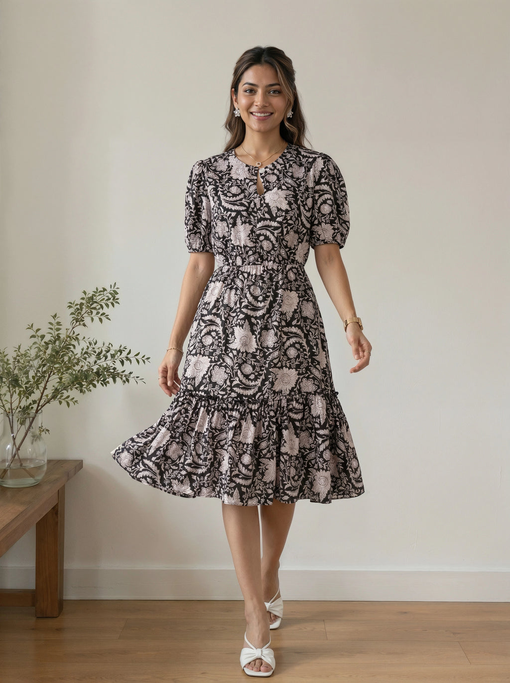 A woman modeling a short-sleeve midi dress with a black and dusty pink floral paisley pattern, standing in a room with a light beige wall and wooden floor.