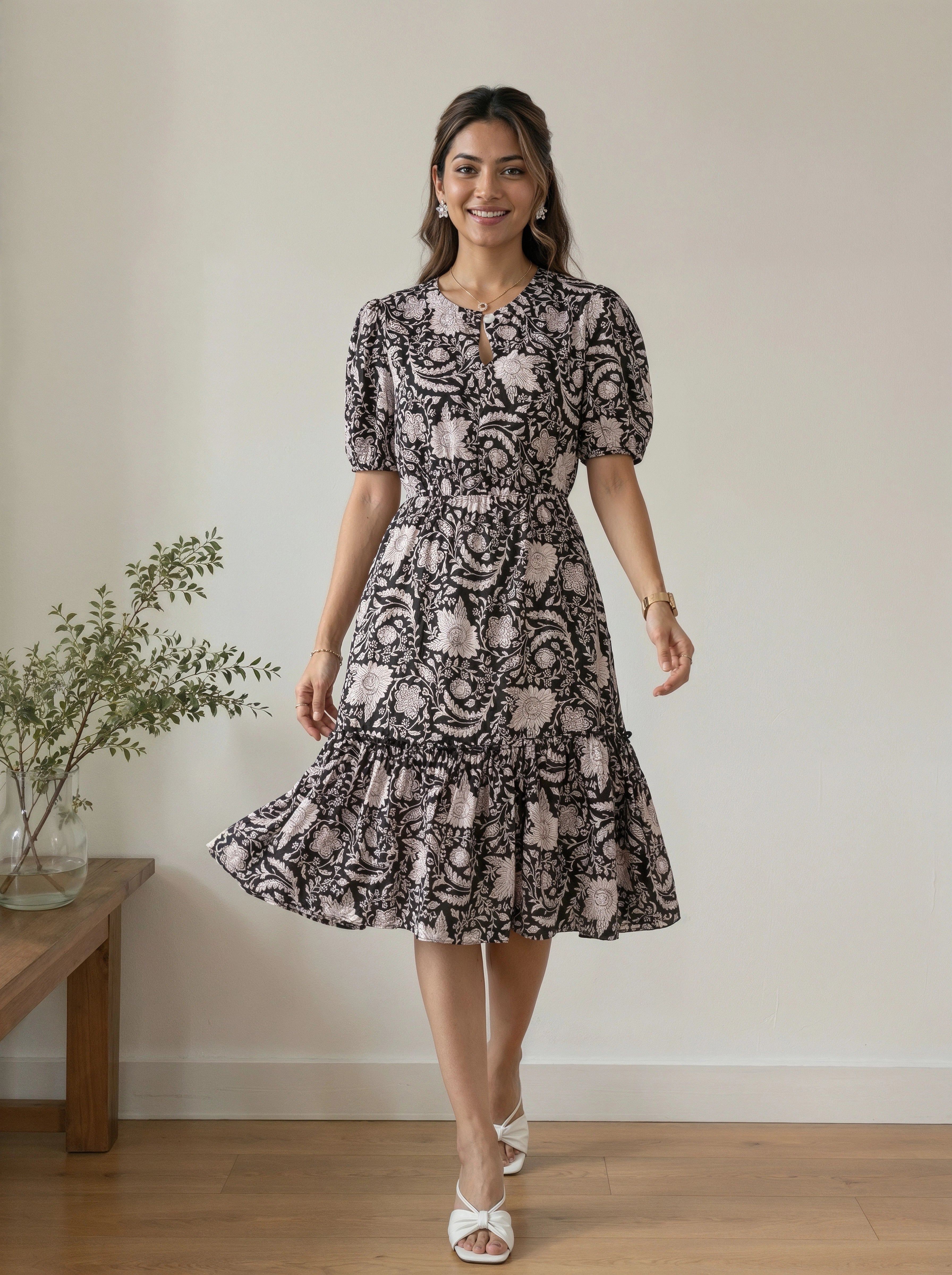 A woman modeling a short-sleeve midi dress with a black and dusty pink floral paisley pattern, standing in a room with a light beige wall and wooden floor.