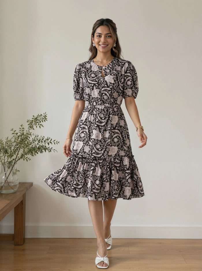 A woman modeling a short-sleeve midi dress with a black and dusty pink floral paisley pattern, standing in a room with a light beige wall and wooden floor.