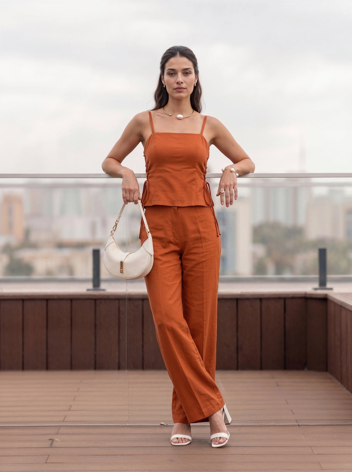 A woman stands on a rooftop terrace wearing a matching rust-orange two-piece set consisting of a sleeveless camisole top and wide-leg trousers. She is holding a white crescent-shaped shoulder bag and wearing white heeled sandals, with a blurred cityscape in the background.