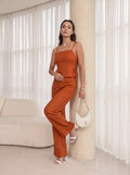 A woman models a matching rust-orange two-piece set consisting of a spaghetti-strap camisole top and wide-leg trousers, styled with a white crescent handbag and white heeled sandals in a bright, modern room.
