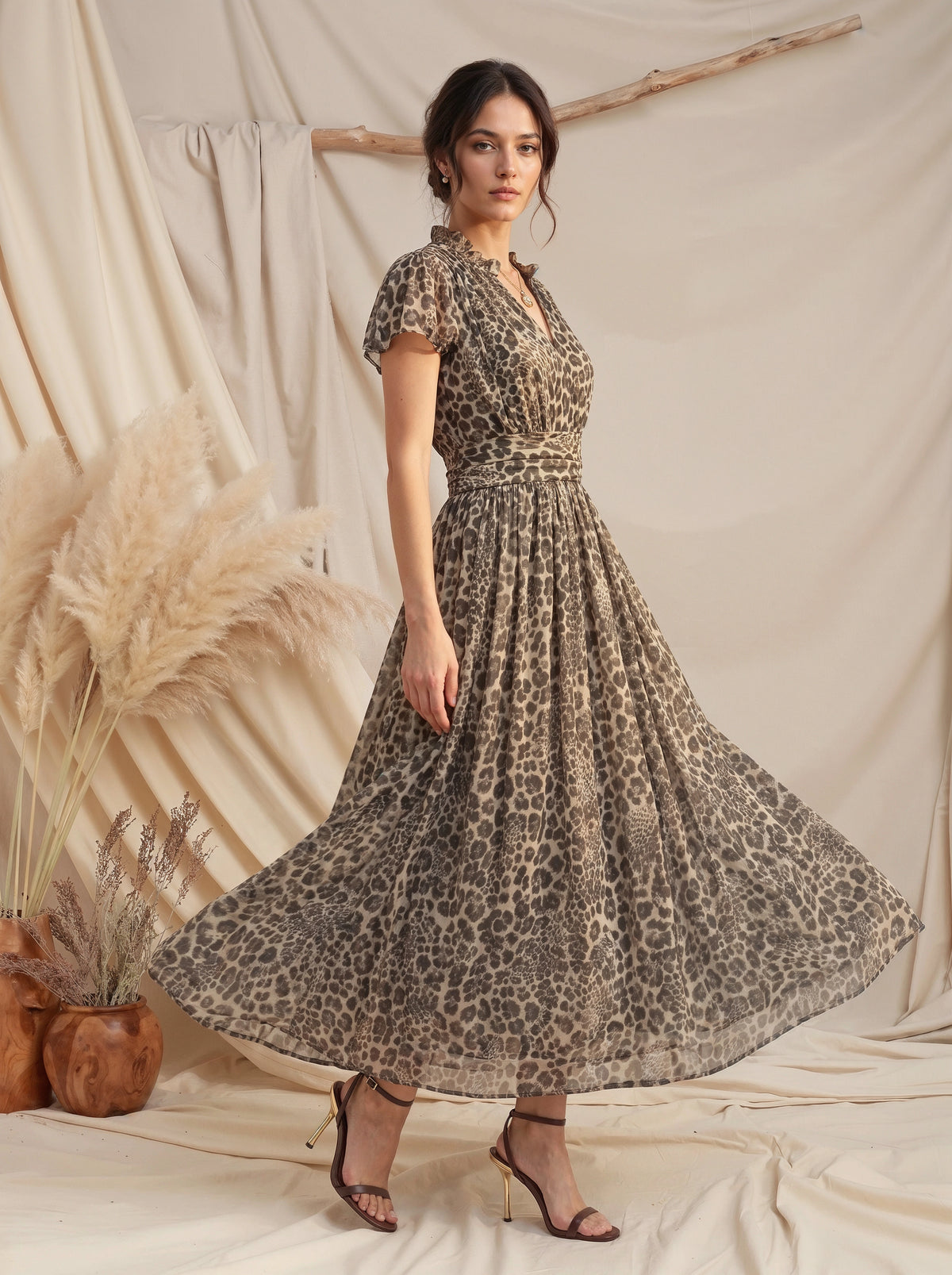 A woman models a leopard-print midi dress with short flutter sleeves, a V-neckline, and a flared skirt, paired with brown strappy heels, set against a draped beige fabric backdrop with pampas grass.