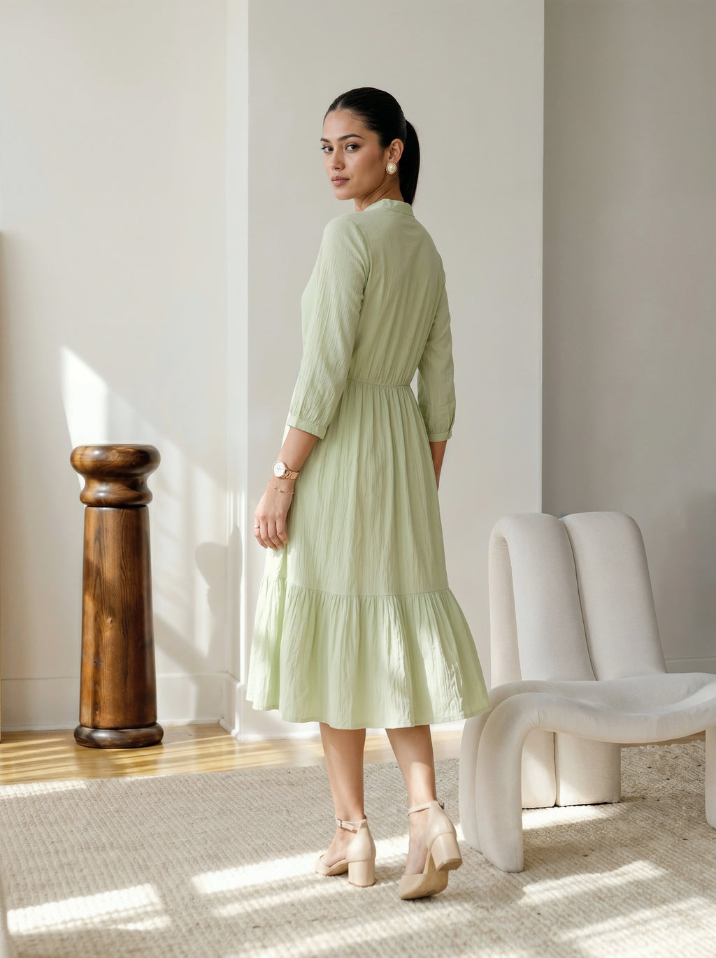 Woman in a light green dress standing in a well-lit room with a white chair and wooden column.