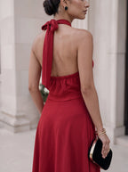 A woman wearing a red halter-neck dress with an open back and a long tie detail, photographed from behind in an outdoor architectural setting.
