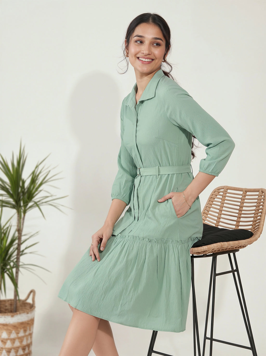 Woman wearing a light green dress standing next to a plant and stool against a white wall.
