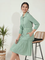 Woman wearing a light green dress standing next to a plant and stool against a white wall.