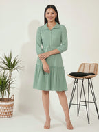 Woman wearing a green dress standing next to a plant and stool on a white background