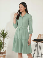 Woman wearing a green dress standing next to a plant and stool against a white wall