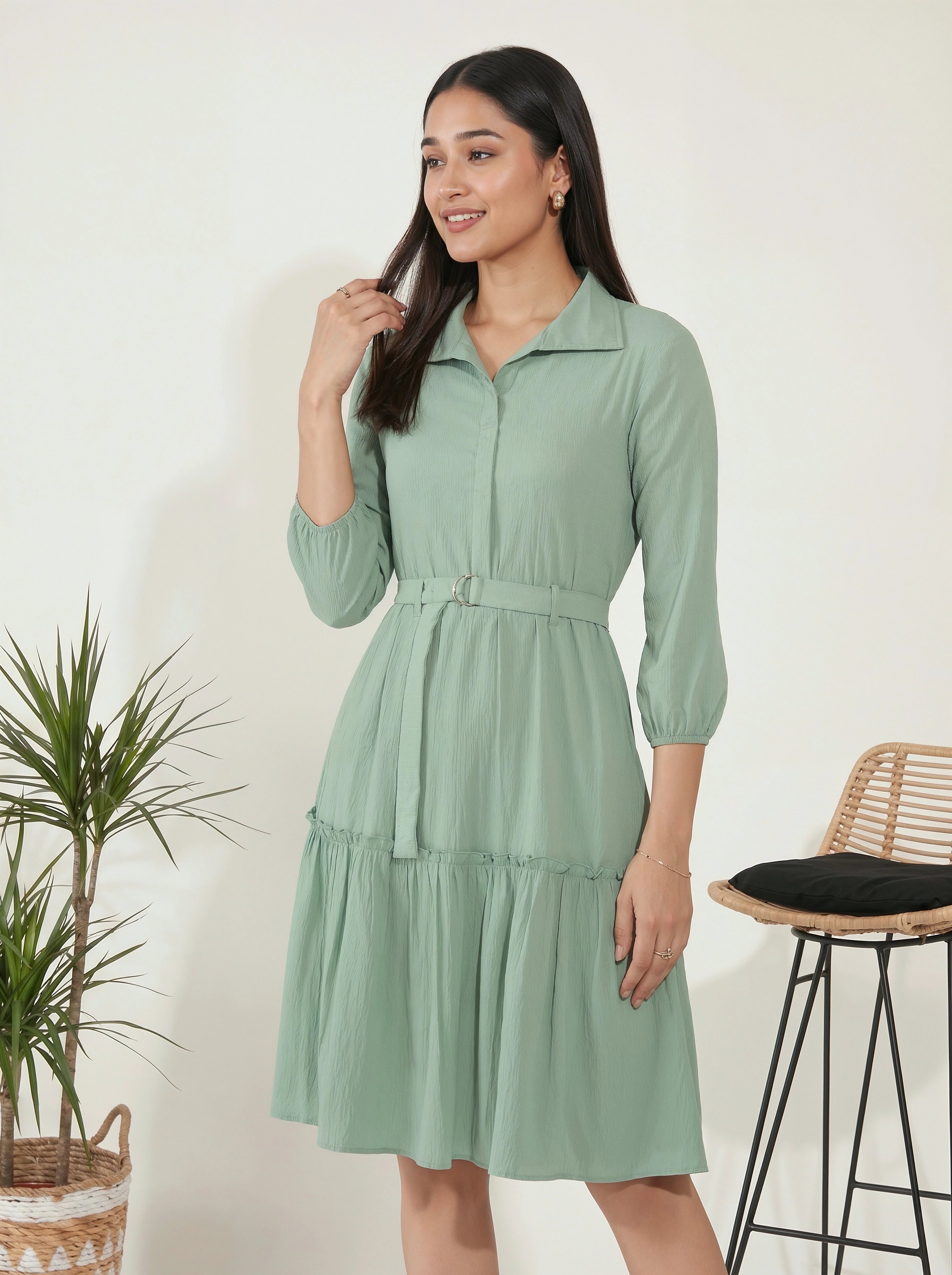 Woman wearing a green dress standing next to a plant and stool against a white wall