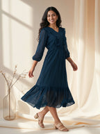 Woman wearing a blue dress standing in a softly lit room with curtains.