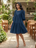 Woman in a blue dress standing in an outdoor setting with plants and tables.